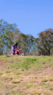Tam teçhizatlı bir motokros binicisi parlak gün ışığında kuru çimenler ve okaliptüs ağaçlarıyla çevrili bir toprak yolda atlar ve yol alır.