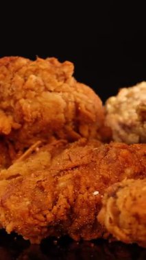 Kızarmış tavuk kanatları, bagetler ve nuggetlar sıcak stüdyo ışıkları altında yansıtıcı bir yüzey üzerinde dönerler ve siyah arka plandaki çıtır çıtır dokuları ortaya çıkarırlar.