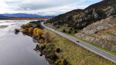 Sonbahar ağaçlarını ve bulutlu gökyüzünün altındaki kayalık tepeleri geçerek, su yansımaları görülebilen bir arabayı takip eden bir dron, dolambaçlı göl yolu üzerinde sorunsuzca uçar.