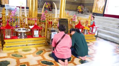 İki kişi Bangkok, Tayland 'da canlı bir tapınakta dua ederken diz çöker. Parlak ışıklandırma dekorasyon ve manevi ortamı vurgular