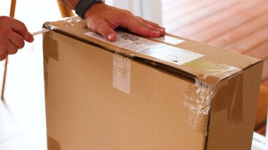 Bir insan dikkatlice bir karton kutuyu açar. Kapalı mekanda iyi aydınlatılmış bir bıçakla.