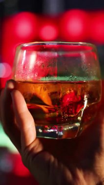 Bir el yavaşça buzlu bir viski bardağını canlı, renkli bir bar arka planına doğru çeviriyor. Ilık ışıklandırma camdaki yoğunluğu ve yansımaları vurgular