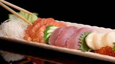Çubuklar, Japon suşi tabağından bir dilim ton balıklı sashimi kaldırır. Stüdyo ışıkları altında, siyah bir arka planda somon, deniz tarağı, yumurta ve garnitür bulunur.