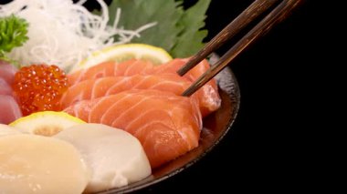 Çubuklar Japon suşi setinden bir dilim somon balığını nazikçe kaldırıyor, parlak, hatta yakın plan çerçeveli, siyah bir zemin üzerine yerleştirilmiş.