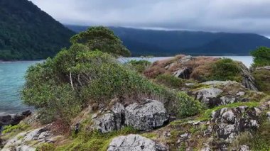 Hava görüntüleri, turkuvaz suları ve bulutlu gökyüzünün altındaki uzak dağlarla kayalık, bitkisel bir kıyı şeridinde süzülerek doğal kıyı manzarasını ve dinamik kamera hareketini vurgular.