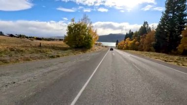 Bir karavan, Yeni Zelanda 'nın Tekapo Gölü' nde sonbahar ağaçları altında ve mavi gökyüzü altında manzaralı kırsal bir yolda ilerliyor. Sabit ileri dönük kamera