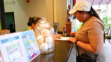 Bir misafir otel resepsiyonuna giriş yapıyor. Parlak, misafirperver bir lobi ortamında çalışanlarla iletişim kuruyor.