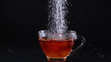 Şeker tanecikleri koyu bir zemine karşı bir bardak çaya dönüşür ve dinamik bir görsel etki yaratır.