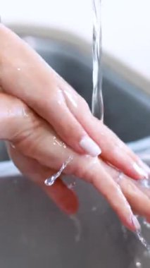 Erişkin ellerin yakın çekimi, açık mutfak lavabosunda akan suyun altında, yumuşak doğal aydınlatma ve sabit kamera odaklı.