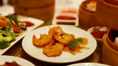 Karides ve sebzeler de dahil olmak üzere çeşitli Dim Sum yemekleri, sıcak ışıkta bir masanın üzerine dizilmiştir.