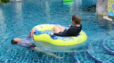 İki insan Yengeç havuzunda güneşli bir günün tadını çıkarıyor. Bir tanesi şişme bir yüzüğün üzerinde yüzüyor, neşeli ve rahat bir atmosfer yaratıyor.