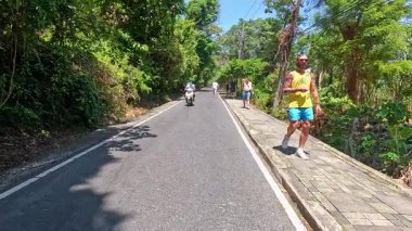Phuket, Tayland 'da turistler yemyeşil ve gezgin scooterlarla çevrili güneşli bir yolda yürüyorlar.