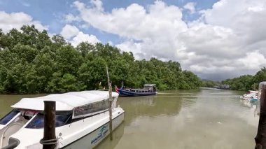 Bir sürat teknesi, parlak gün ışığında sakin bir mangrov nehri boyunca, Ko Phayam 'daki yemyeşil ve parçalı bulutlu gökyüzü ile ahşap bir rıhtıma yaklaşıyor.