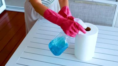 Pembe eldiven giyen biri, kapalı bir ortamda beyaz bir masanın üzerine mavi temizlik sıvısı sıkıyor. Sabit bir yukarıdan aydınlatma ve statik kamera açısı kullanıyor.