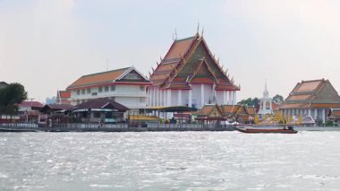 Geleneksel Tayland mimarisi ve yumuşak gün ışığında teknelerle sakin bir nehir manzarası.