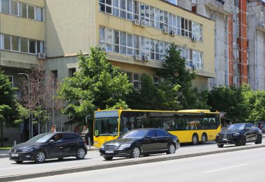 Kosova, Priştine-27 Mayıs 2025: Yolcularla dolu Sarı Kamu Otobüsü, yürüyen ve ayakta duran insanlar, Trafik Yoğun Saati sırasında Parlak Siyah Arabalar ve Yoğun Trafik