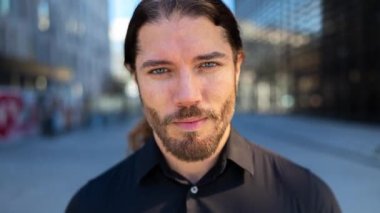 Arka planda ofis binaları olan ve kameraya yaslanan genç bir adam.