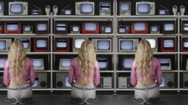 Arızalı televizyon ekranlarından oluşan duvarı arkadan izleyen bir kadın. 