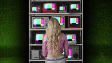 Arızalı televizyon ekranlarından oluşan duvarı arkadan izleyen bir kadın. 
