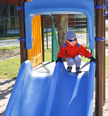 Ceketli ve şapkalı küçük çocuk baharda kaydırağın üstünde oynuyor.