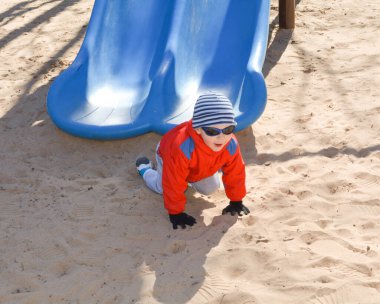 Ceketli ve şapkalı küçük çocuk baharın başında kaydırağın üzerinde oynuyor.