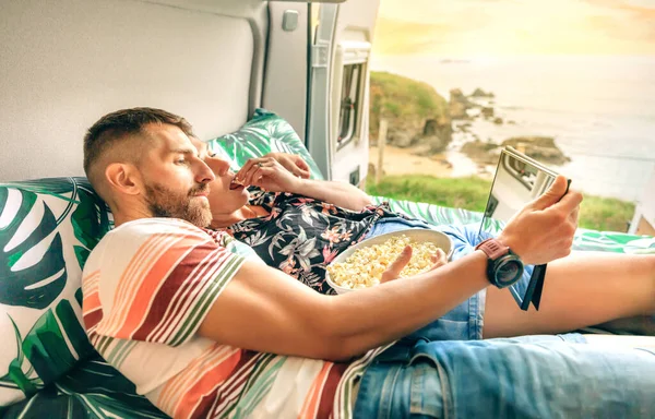 若いですカップル見ますAムービーオンザタブレット食べるポップコーン嘘のベッドの上に彼らのキャンパーバンでドアを開けて海岸の風景に向かって — ストック写真