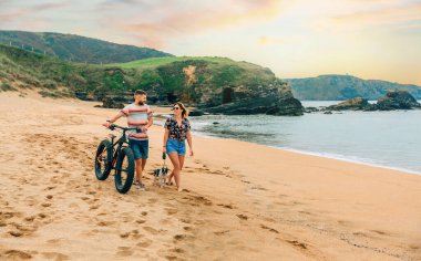 Couple with a fat bike taking a walk on the beach with their dog