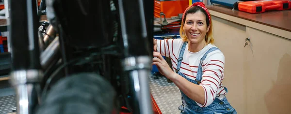工場でオートバイを修復しながら カメラを見てそばかすと笑みを浮かべて整備士の女性の肖像画 — ストック写真