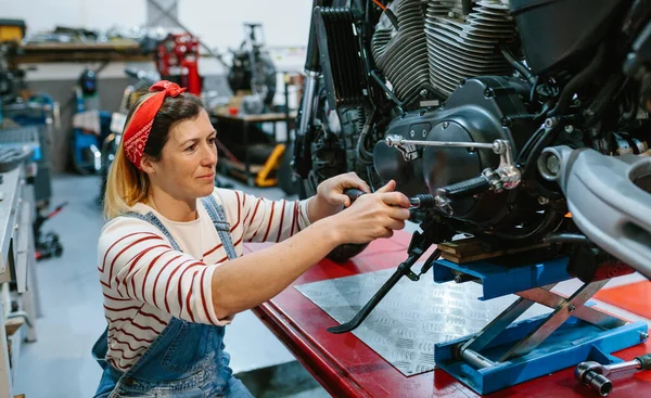 工場上のプラットフォーム上のカスタムバイクのエンジンをレビューツールと幸せな整備士の女性の肖像 — ストック写真