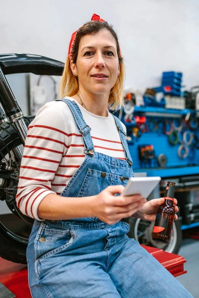 工厂里 坐在摩托车前拿着手机和啤酒瓶看着相机微笑的机械师妇女的画像 — 图库照片