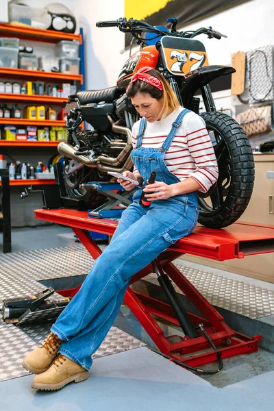 快乐的机修工女人一边看着电话 一边拿着一瓶啤酒坐在平台上 一边在工厂里量身定制摩托车 — 图库照片