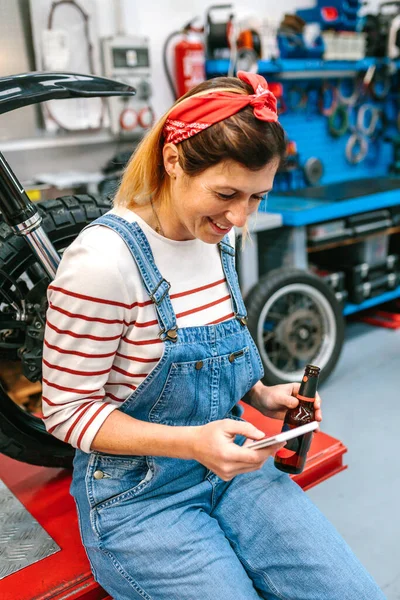 工厂里坐在摩托车前 面带微笑的机械师女人拿着一瓶啤酒看着电话的画像 — 图库照片