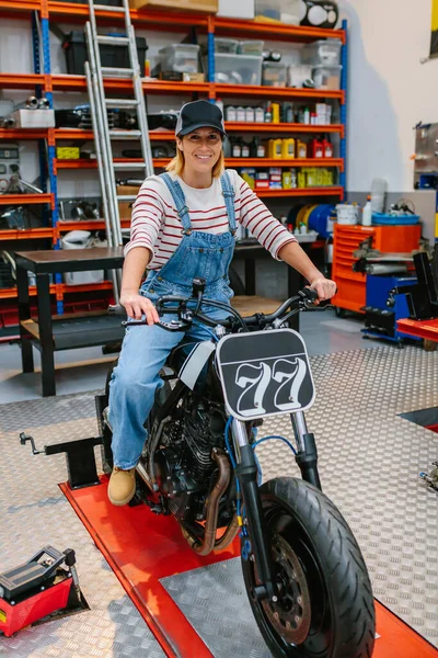 Portret Szczęśliwej Mechaniczki Czapką Patrzącej Kamerę Siedzącą Nad Niestandardowym Motocyklem — Zdjęcie stockowe