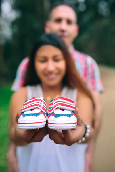 微笑的拉丁裔孕妇露出小婴儿运动鞋 而她的伴侣则以她的腹部为背景 有选择地关注未来的运动鞋 — 图库照片