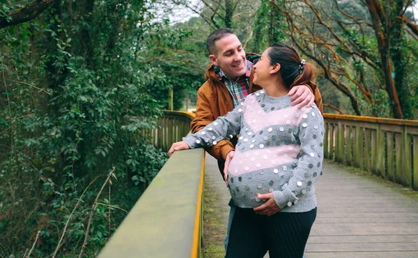 笑笑的高加索男性期待着快乐的拉丁裔孕妇抱着她的肚子站在大自然的小径上 — 图库照片