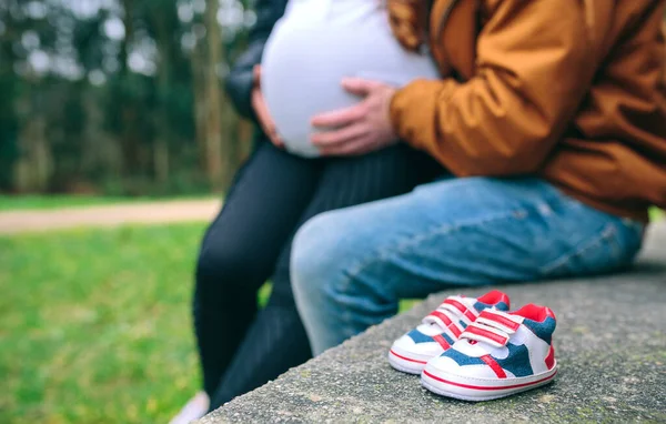 在长椅上的小婴儿运动鞋和无法辨认的怀孕女性和伴侣在背景下抱着她的腹部 有选择地关注未来的运动鞋 — 图库照片