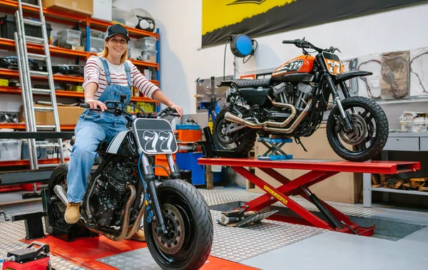 Szczęśliwy Mechanik Kobieta Czapką Patrząc Aparat Siedzi Własny Motocykl Fabryce — Zdjęcie stockowe