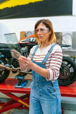 Güvenlik gözlüklü kadın tamirci fabrikadaki motosikleti incelemek için dijital tablete ve tanı uygulamasına dokunuyor.