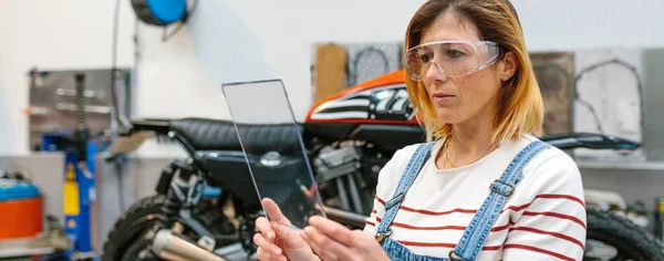 Soustředěná Mechanička Bezpečnostními Brýlemi Průhledným Tabletem Před Motocyklem Garáži — Stock fotografie