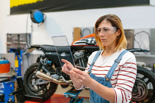 Soustředěná Mechanička Bezpečnostními Brýlemi Dotýkající Průhledného Tabletu Před Motocyklem Garáži — Stock fotografie