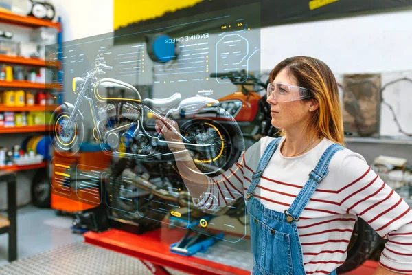 工場でオートバイを確認するために拡張現実のホログラムとハードパネル画面に触れる眼鏡を持つ若い女性のメカニック 未来的なユーザーインターフェイスの概念 — ストック写真