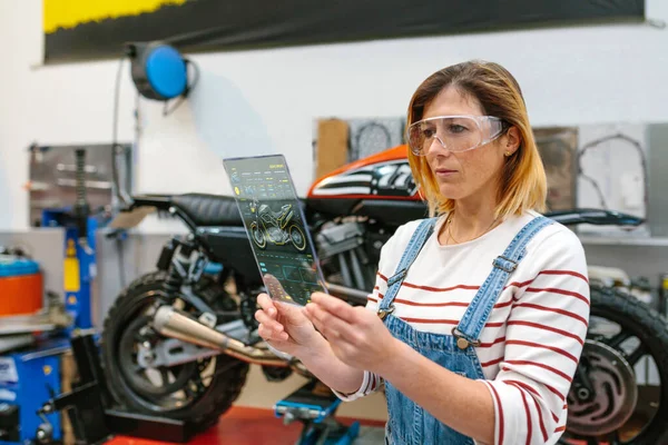 利用透明数码平板和诊断应用程序对工厂摩托车进行审查 集中配备安全眼镜的女技师 — 图库照片