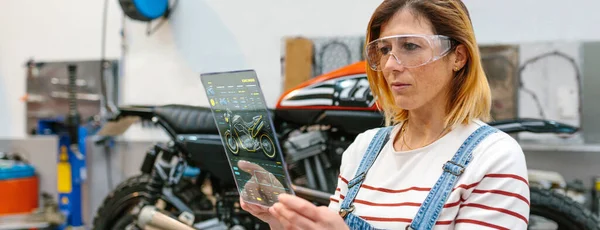 聚精会神的女机修工 戴着安全眼镜 触摸透明的数码平板电脑 配有诊断应用程序 可在工厂查看摩托车 — 图库照片
