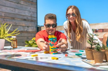 Güneş gözlüklü genç adam, yaz partisinde çatıdaki kız arkadaşının yanına Jenga oyunu parçası koyuyor. Jenga kulesi oyununa seçmeli odaklan.