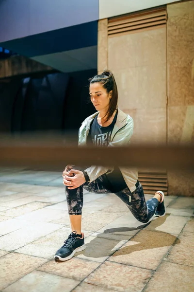 空の町で夜に訓練する前に彼女の足を伸ばすポニーテールと集中した若いブルネットの女性ランナー — ストック写真