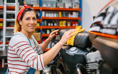 Fabrikada mikrofiber bezle motosiklet koltuğunu temizlerken kameraya bakan çilli ve saç bandanalı kadın tamircinin portresi