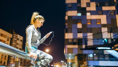 Konsantre genç sarışın kadın akıllı telefon uygulamasında canlı yayın eğitimi videosuna bakıyor. Kulaklıklı genç bayan koşucu gece vakti tırabzanın üzerinde oturan müziği dinliyor. Sağ kopyalama alanı.