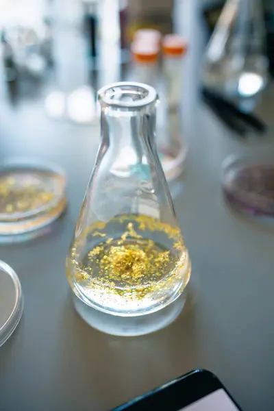 Close up of erlenmeyer flask with golden glitter sample mixed in analysis liquid over a table on environment research lab. Study of dangerous microplastics behavior to humans and environment concept.