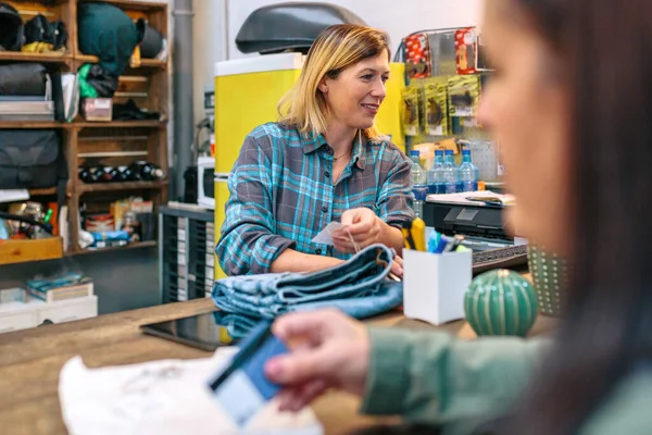 元気な若い女性従業員がコンピュータでジーンズの価格をチェックしながら 顧客は現地の店舗で購入するクレジットカードを持っています 女性ショップアシスタント クライアントに衣料品を現金化 — ストック写真