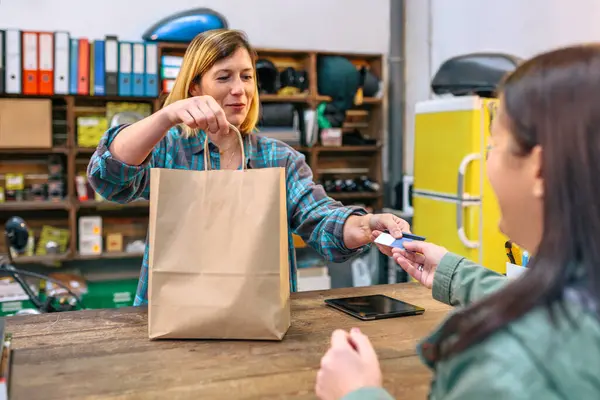 笑顔のブロンドの若い女性ショップアシスタントは 購入した衣料品を女性のお客様にクラフト紙袋を届けます 顧客のクレジット カードを取って店で購入を満たす幸せな女性の従業員 — ストック写真